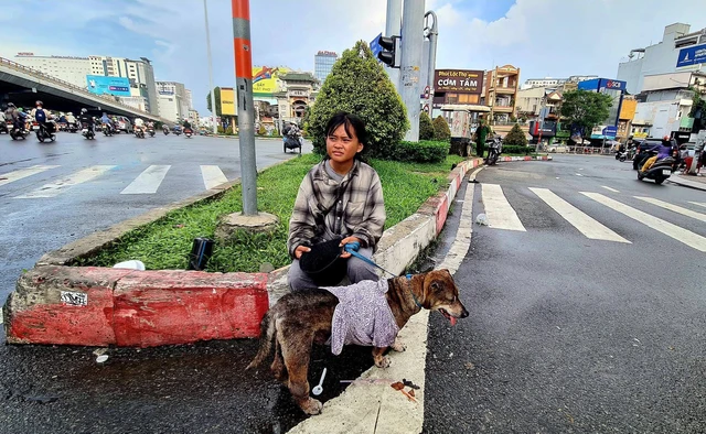 Ngồi nép bên vỉa hè tại giao lộ Hàng Xanh, cô bé đội chiếc mũ cũ sờn màu, áo sơ mi bạc phếch phủ kín đầu gối.